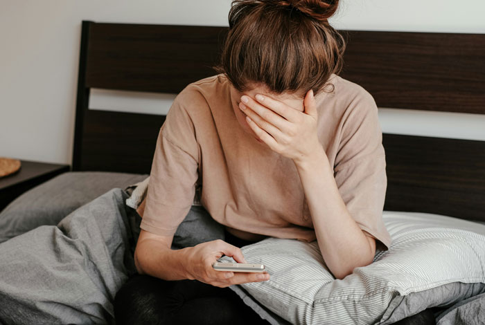 Woman sitting on bed, looking distressed, holding a phone, relating to husband denies leaving children home alone.