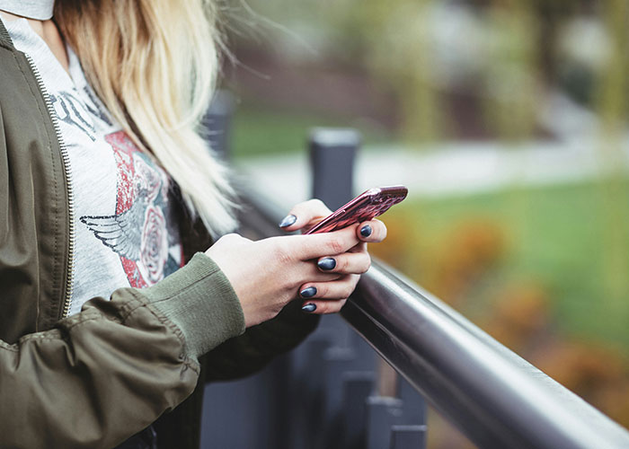 Person with painted nails holding a phone by a railing, contemplating tattoos before getting inked.