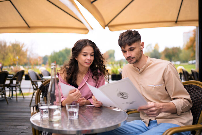 A couple on a first date at an outdoor cafe, discussing the menu while sitting under umbrellas.