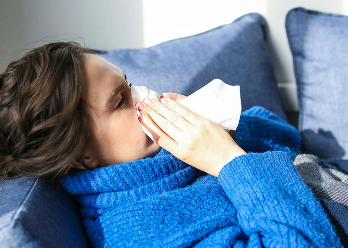 Person in a blue sweater lying on a couch, using a tissue.