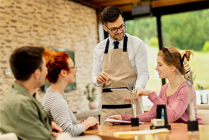 People sitting at a restaurant table, a waiter taking their order, meal payment discussed among friends.