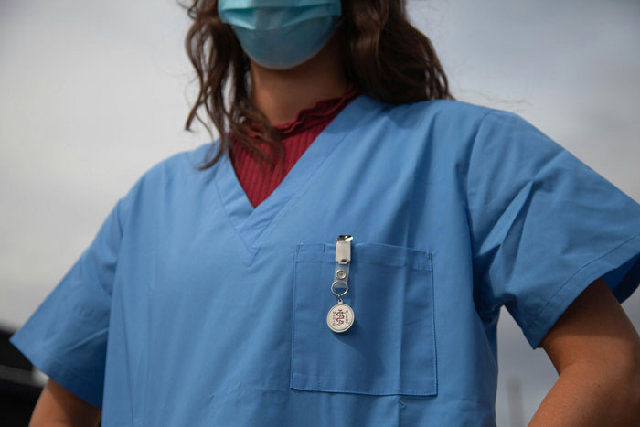 Woman in blue nursing scrubs, wearing a mask, standing confidently under a clear sky.