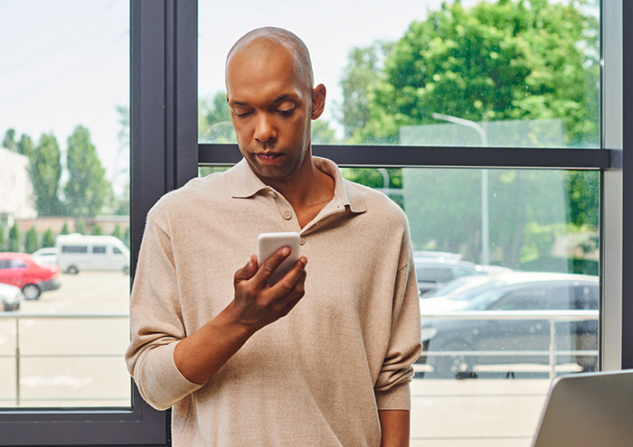 Man looking at his phone, decision impacting marriage, in a bright room with a view of parked cars and trees.