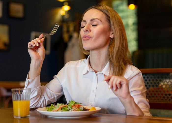 Woman dining at a restaurant table, enjoying her meal with a fork, a glass of orange juice beside her.