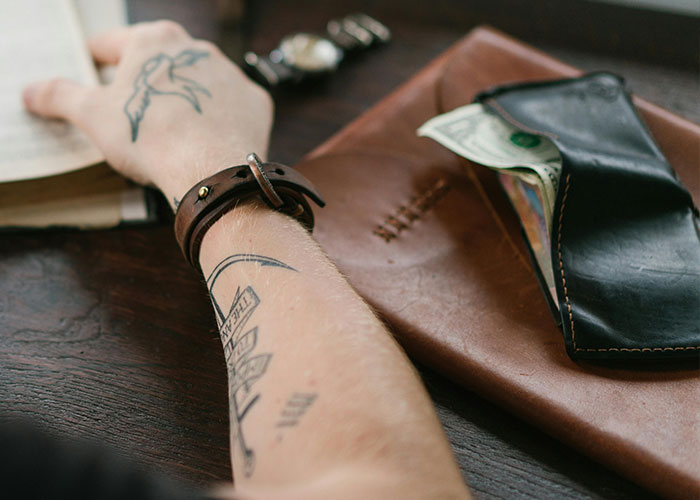 Arm with tattoos resting on a table next to a leather wallet and book.