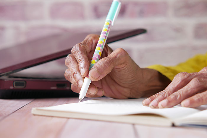 Older woman writing with a colorful pen near a laptop, contemplating inheritance decisions.