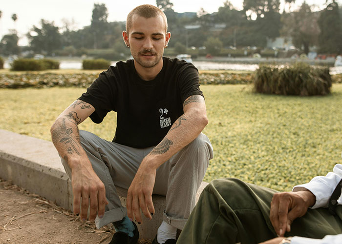 Man with tattoos sitting outdoors in casual attire, showcasing inked designs on arms.