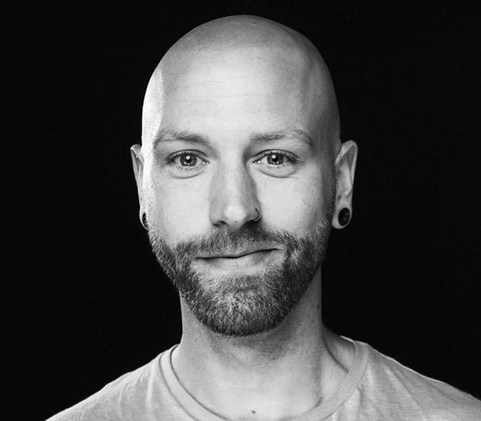 Bald man with a beard wearing earrings, neutral expression, against a dark background, discussing Covid vaccines.