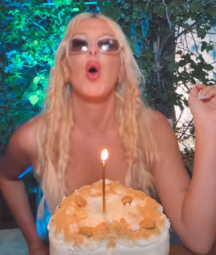 Blonde woman celebrating with a lit birthday candle on a cake, wearing sunglasses, greenery in the background.