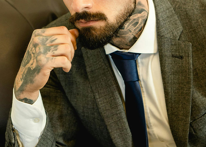 Man in a suit with hand tattoos and a beard, contemplating decisions before getting inked.