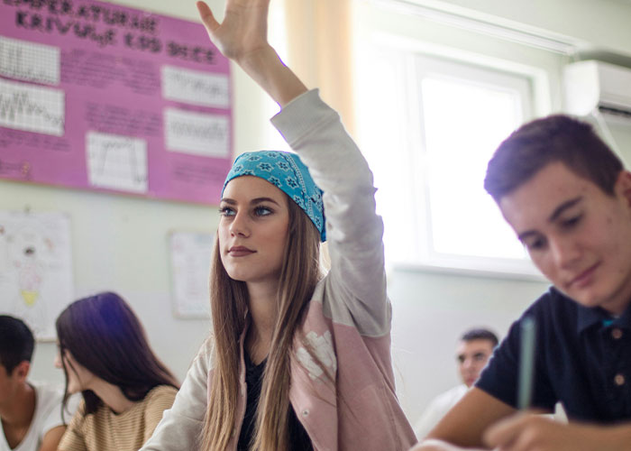 Estudiante con pañuelo azul en clase, levantando la mano, relacionada con excusas de estudiantes que fueron ciertas.