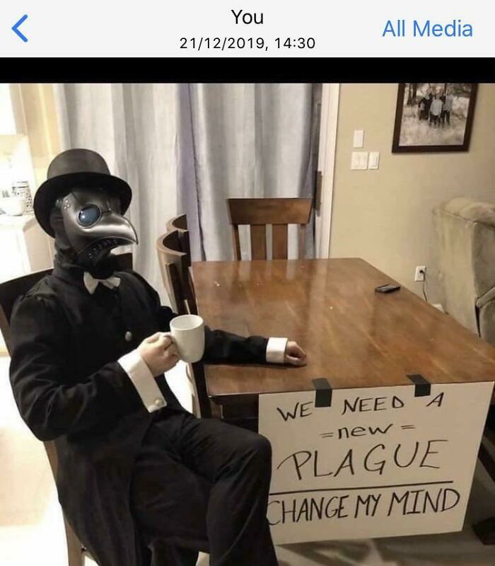 Person in plague doctor costume sitting at table with humorous sign, reflecting on bad future predictions.