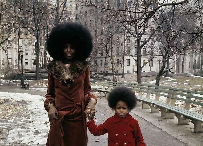 A person and child wearing red coats with afros in a winter urban park, reflecting rare historical moments.