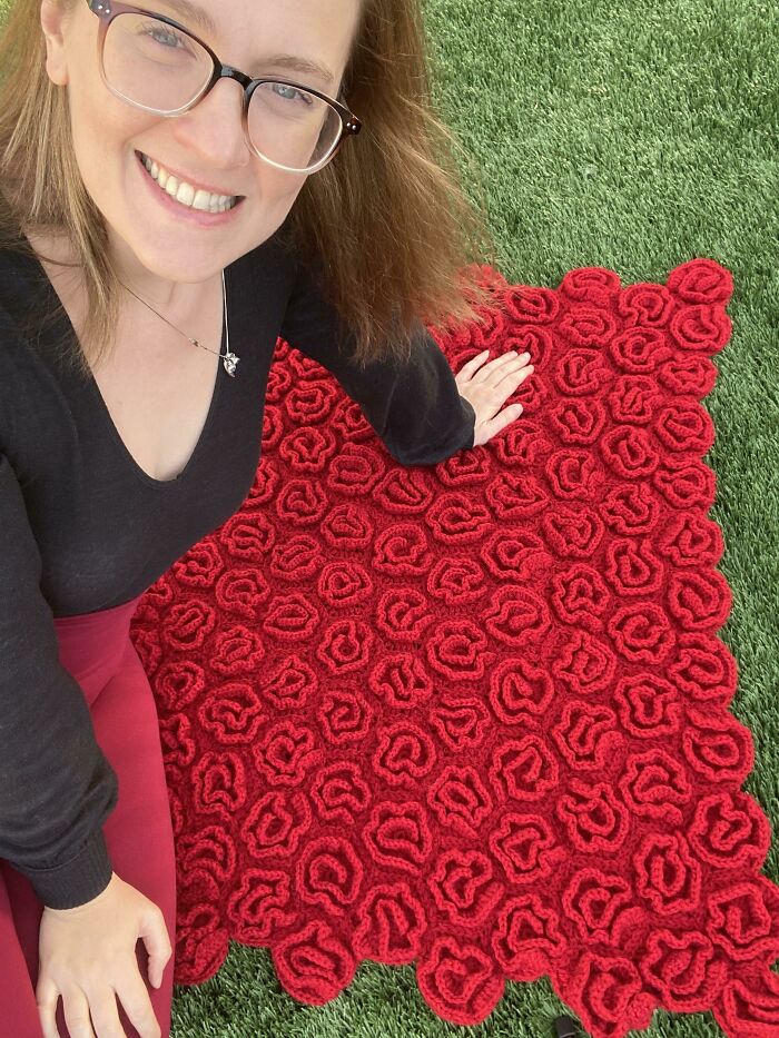 Woman smiling while displaying a red, rose-themed crochet piece on grass, ideal Valentine’s Day gift inspiration.