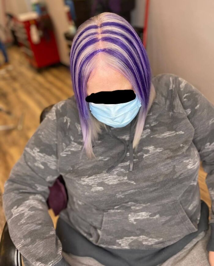 Person with a purple and white striped hair fail, wearing a face mask and camouflage hoodie, sitting in a salon chair.