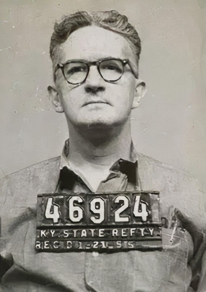 A man with glasses in a vintage mugshot, wearing a collared shirt, holding a numbered sign.