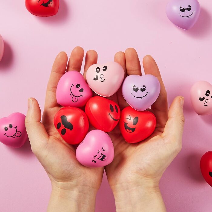 Hands holding playful, heart-shaped Valentine's Day gifts with cute faces on a pink background.