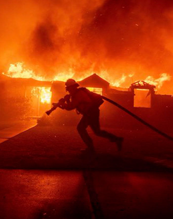 Firefighter battling intense flames with hose amidst a house fire at night.