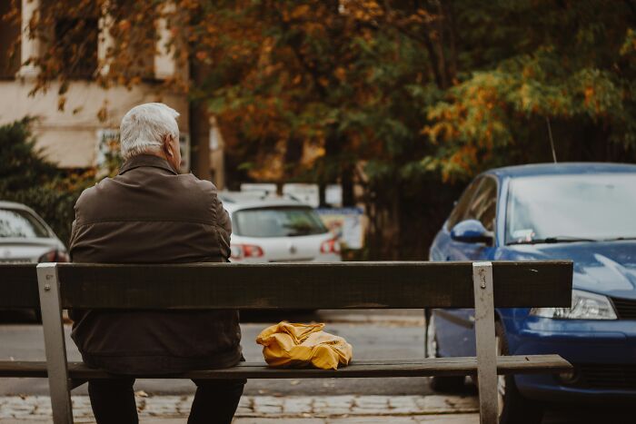 Elderly man on a park bench with a yellow bag, cars and autumn trees in background.