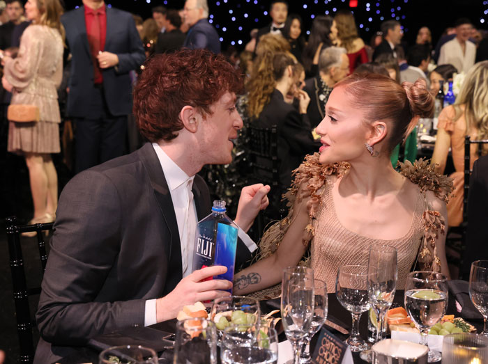 Ariana Grande at Critics Choice Awards seated at a table, engaging in conversation with a man holding a water bottle.