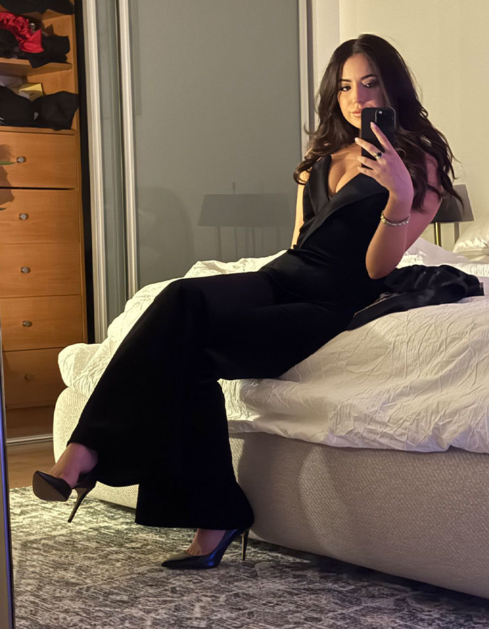 Woman in a black outfit seated on a bed, taking a mirror selfie, related to Elon Musk's 13th child news.