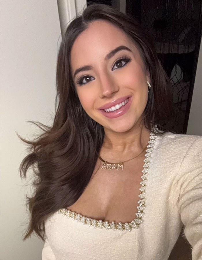 Smiling woman with long hair in a square-neck top, wearing a name necklace.