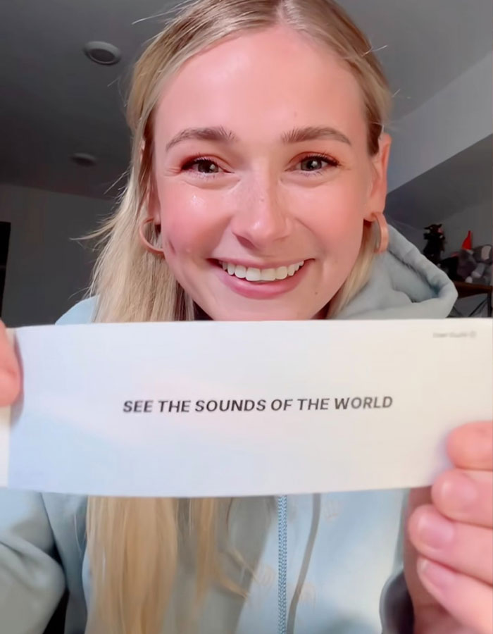 Smiling woman holding a card that reads "SEE THE SOUNDS OF THE WORLD.