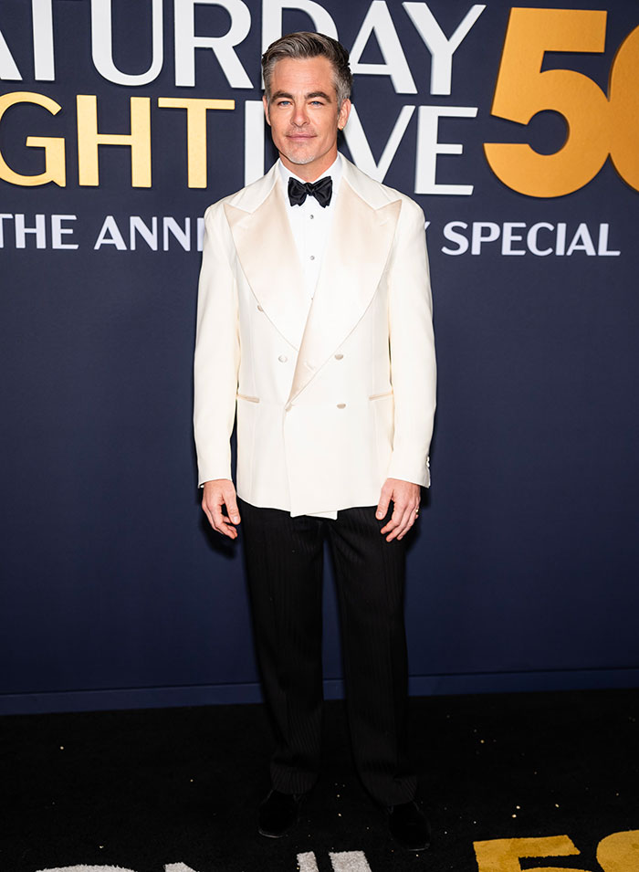 Man in a white tuxedo with a black bow tie on SNL 50th red carpet, posing against event backdrop.