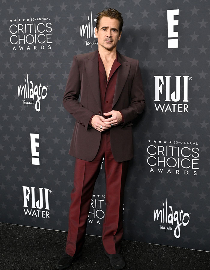 Man in a burgundy suit at Critics Choice Awards red carpet, standing against a backdrop with event logos.