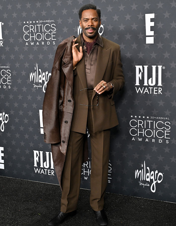 Man in brown suit with coat and glasses at Critics Choice Awards red carpet.