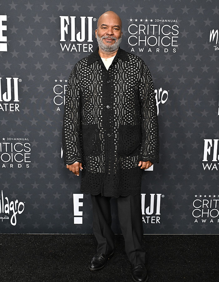 Man in a daring outfit at the Critics Choice Awards red carpet, wearing a patterned black coat and black pants.