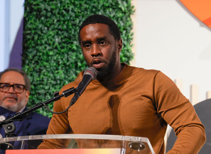 Man speaking into a microphone at an event, wearing a brown shirt, with Diddy's lawyer in the background.