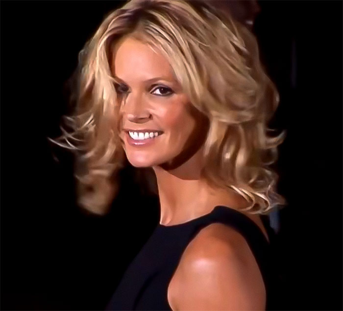 I don't know who this is. Person smiling with blonde hair, wearing a black dress, facing sideways against a dark background.