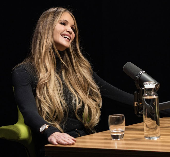 Person with long hair in black outfit speaking at a microphone, related to heavy photoshop discussion.