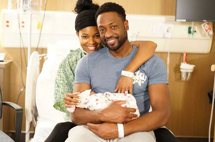 Couple smiling while seated in a hospital room, holding a newborn in their arms, highlighting surrogate family moments.