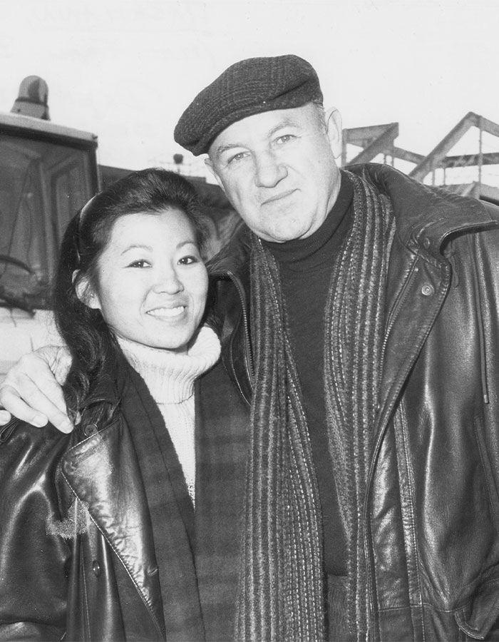 Two people in winter clothing, smiling, with a backdrop of a snowy outdoor scene, related to Gene Hackman investigators.