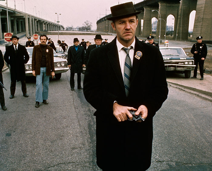 A man in a black coat and hat stands on a street with others and police cars behind him.