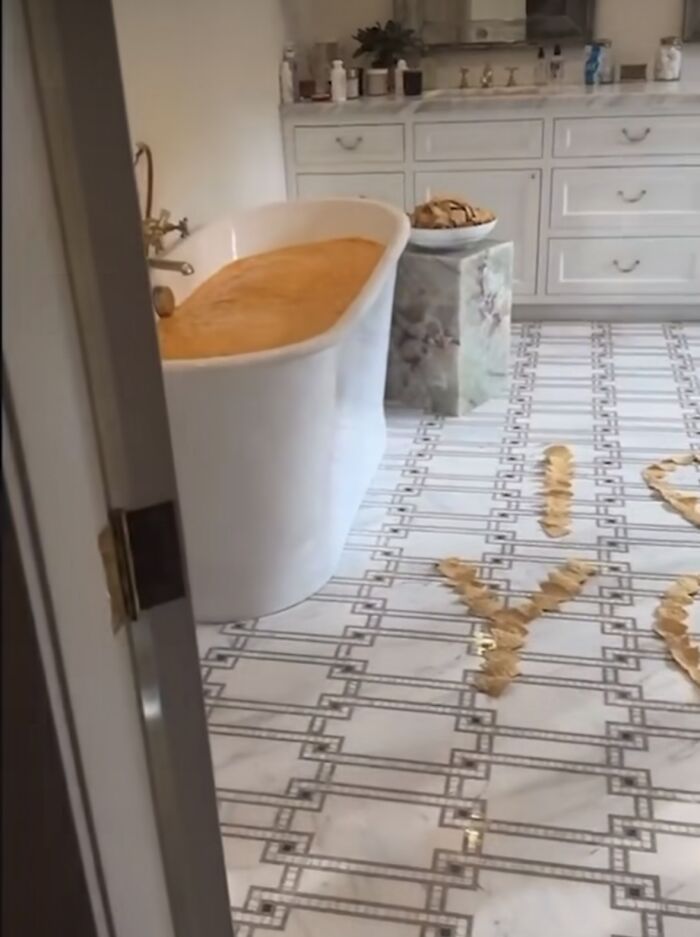 Bathtub filled with orange water, cookies on a pedestal, and "I Love You" spelled with objects on the floor.