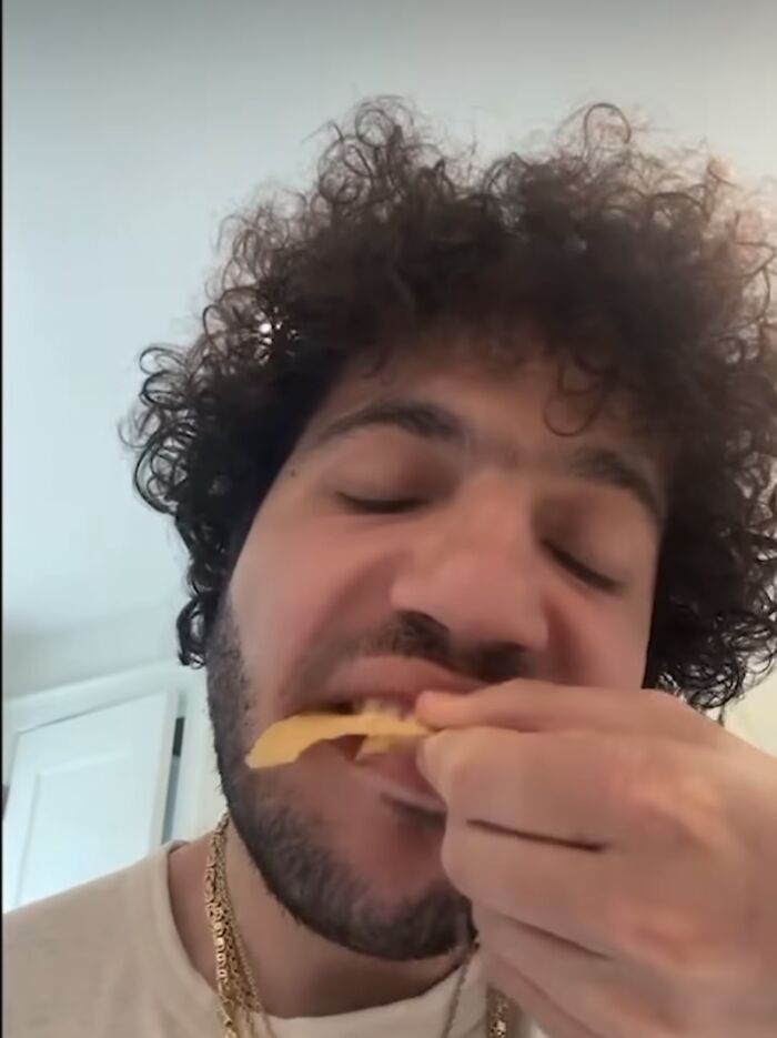 A person with curly hair biting into a chip, wearing a white shirt and gold chains, related to Valentine's Day gift topic.