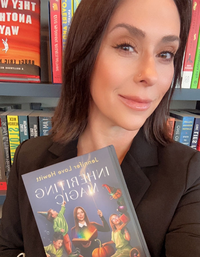 Woman holding a book titled "Inheriting Magic" in front of a bookshelf, highlighting a bare-faced look and natural aging.