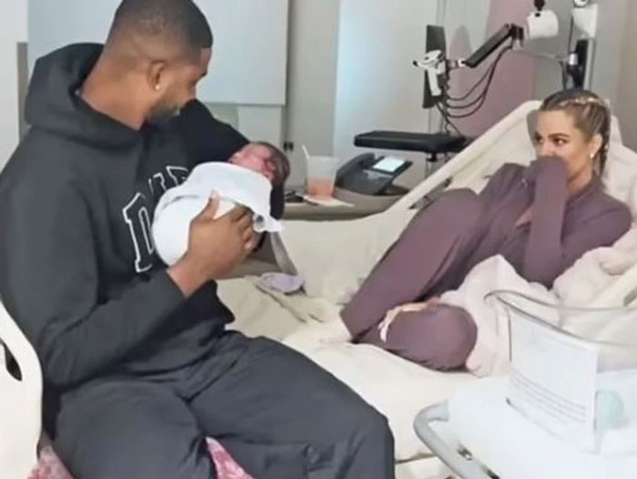 Celebrity couple in hospital room with surrogate baby, seated on bed, sharing a tender moment.