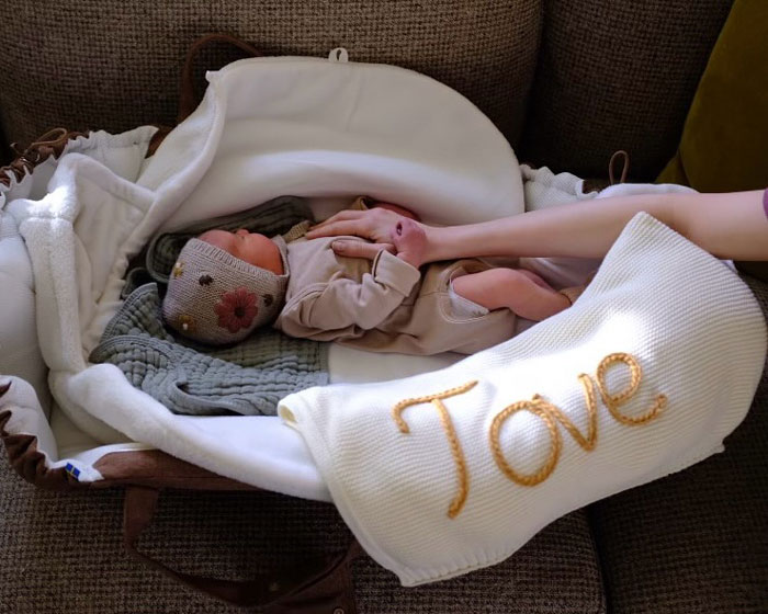 Newborn baby in a cozy basket labeled "Tove," held by an unseen person, symbolizing surrogate journey.