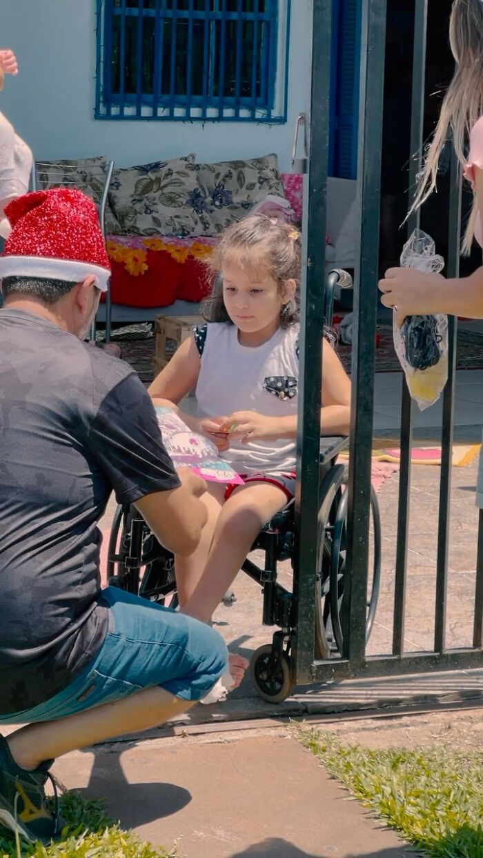 Man in Santa hat kneeling by a young girl in a wheelchair near a gate, handing her a book.