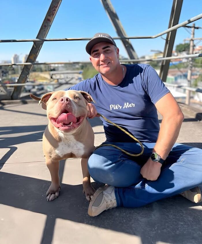 Man sitting with a happy pit bull on a rooftop, wearing a blue shirt and cap.