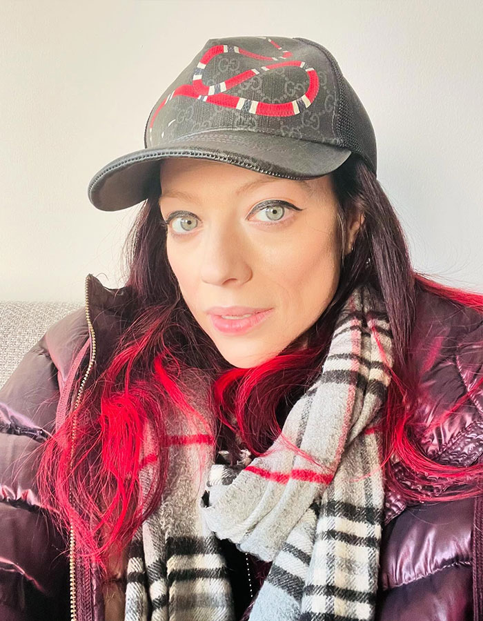 Woman with a hat and red-tinted hair, wearing a patterned scarf, smiling softly at the camera.