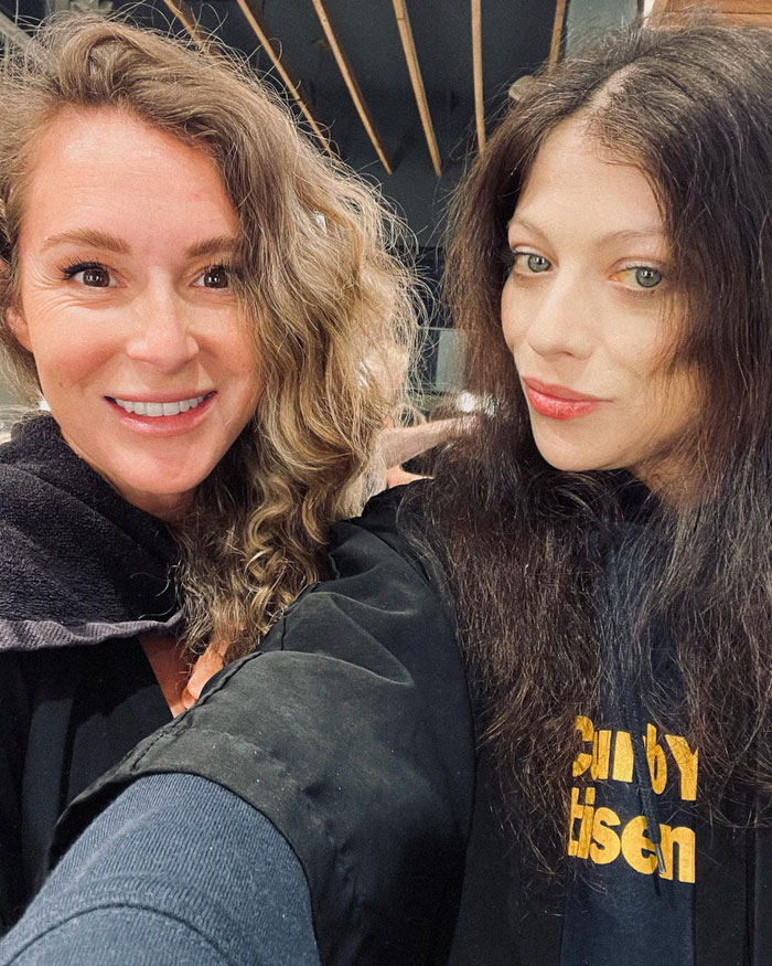 Two women smiling, one with curly hair, the other with straight hair, casually posing indoors.