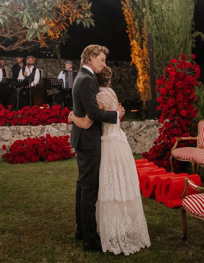 Millie Bobby Brown hugging someone at an event, surrounded by red roses and greenery.