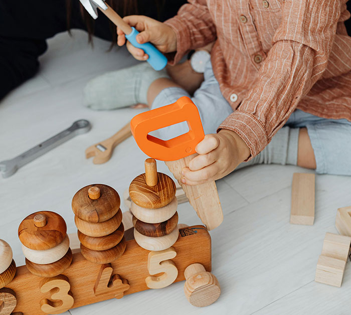 Child playing with wooden toys on the floor, holding an orange toy saw, symbolizing birthday party preparations.