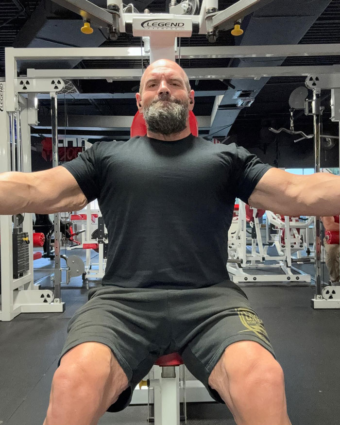 Man in a gym seated on exercise equipment, arms extended, wearing a black shirt, reflecting on drastic weight loss.