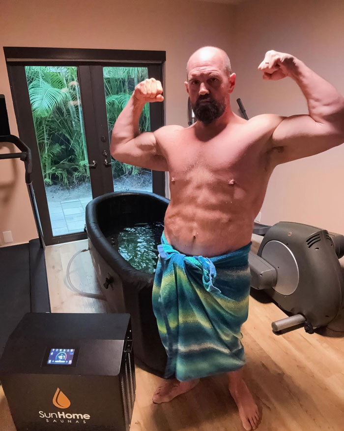 Man flexing muscles post-300-lb weight loss, using Ozempic, in home gym with towel and sauna machine.